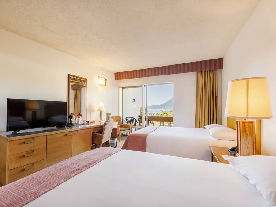 A hotel room with two beds, a desk with a TV, a chair, a lamp, and a view of the outdoors.