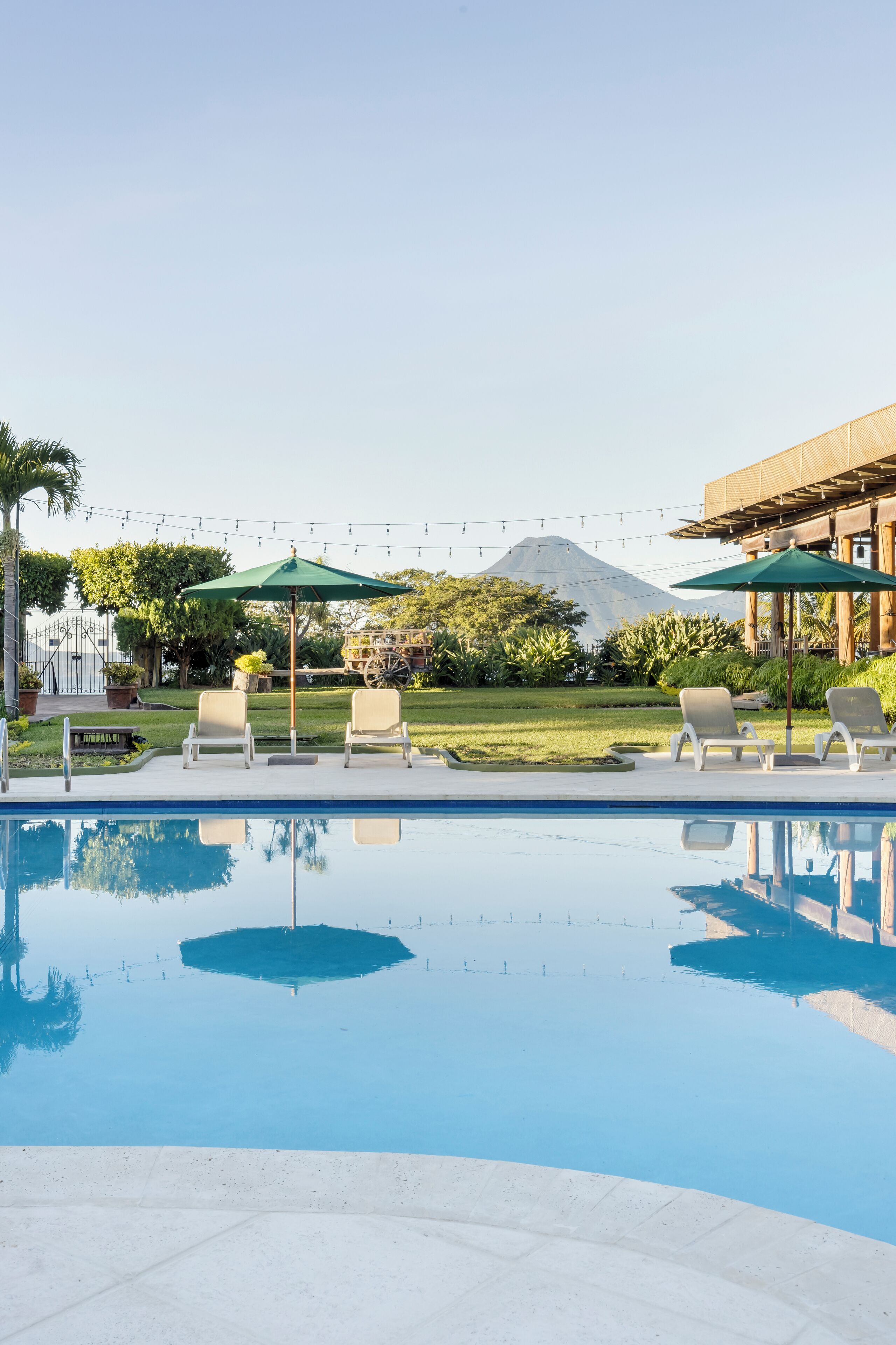 A swimming pool with lounge chairs and umbrellas, surrounded by greenery and a building with a mountain in the background.