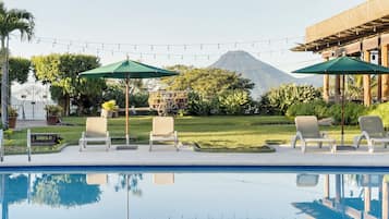 A swimming pool with lounge chairs and umbrellas, surrounded by greenery and a building with a mountain in the background.