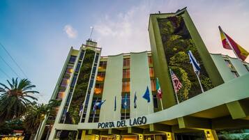 Un hotel llamado "Porta del Lago" con varias banderas, balcones con vegetación y un cielo despejado.