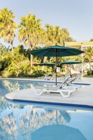 Una piscina con sillas de descanso y sombrillas, rodeada de palmeras y un edificio al fondo.