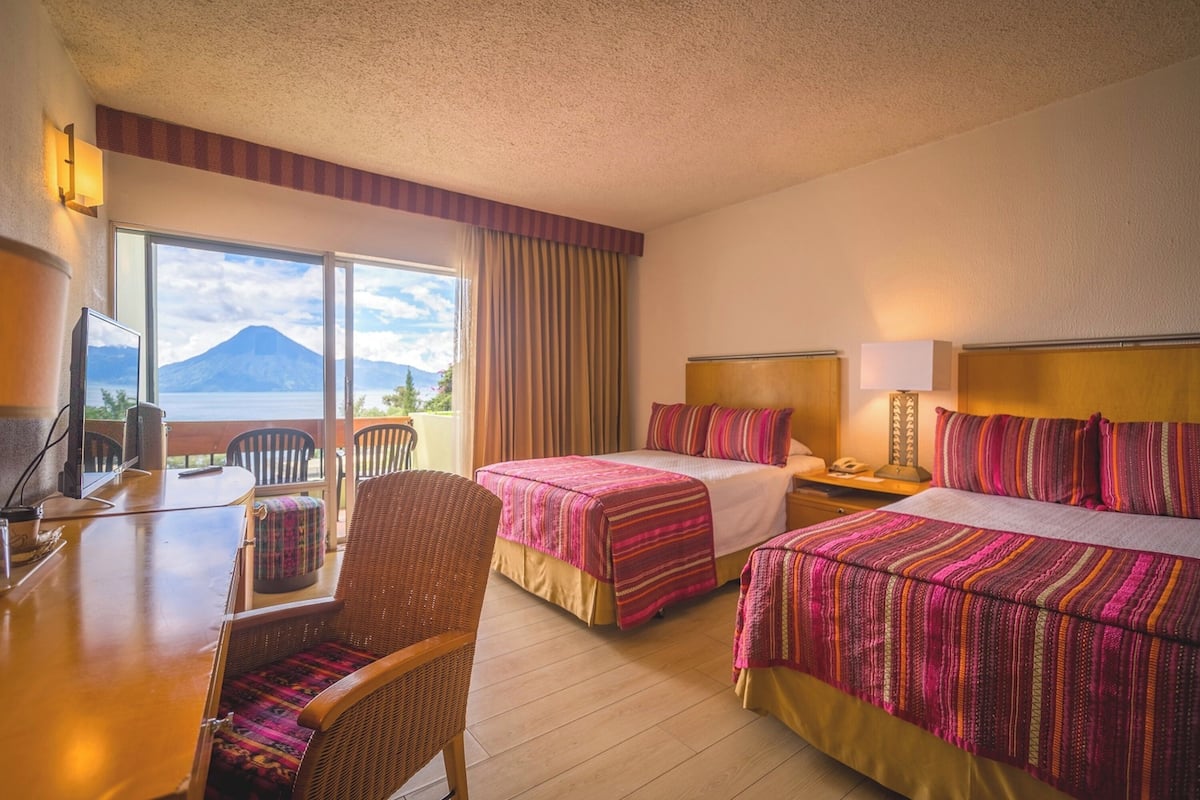 A hotel room with two beds, a desk, a chair, and a view of a mountain.