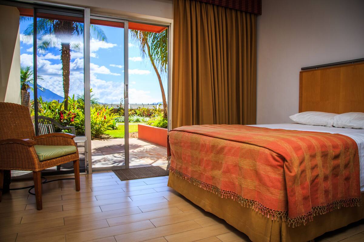 A hotel room with a bed, a chair, and a view of a garden and mountains through a sliding door.