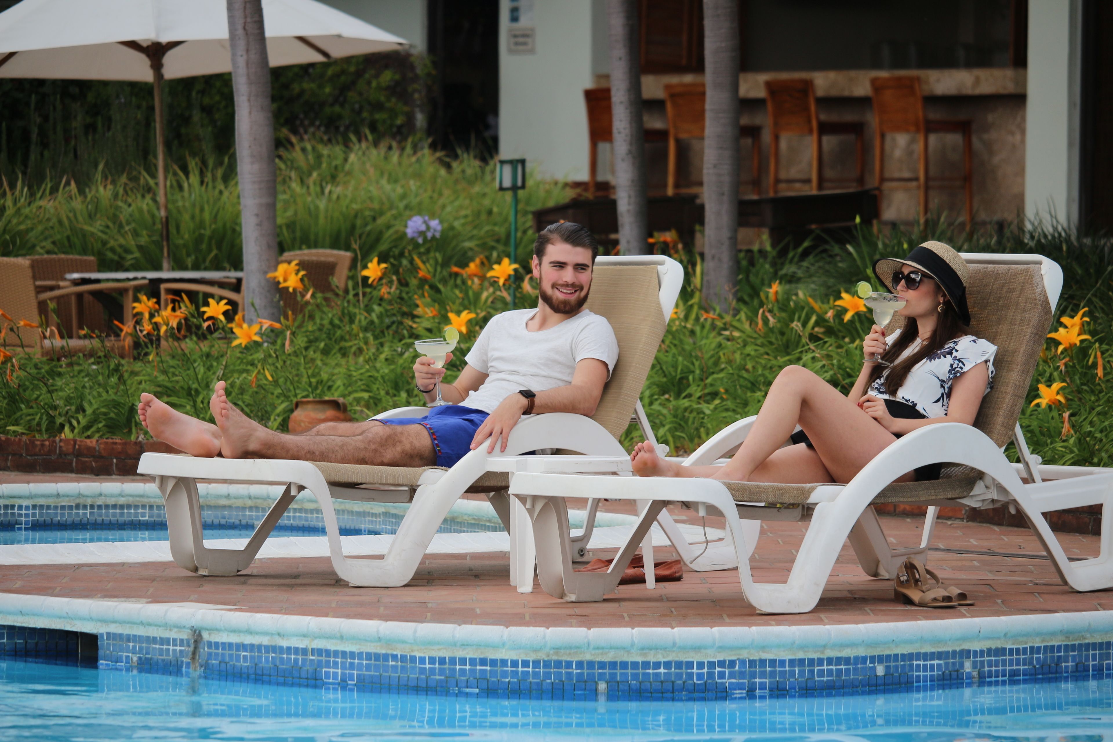 Dos personas descansando junto a una tumbona junto a una piscina con azulejos y muebles de jardín al fondo.