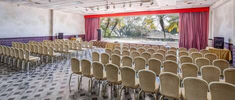 A conference room with rows of wicker chairs, a stage with a red curtain, and a view of palm trees through large windows.