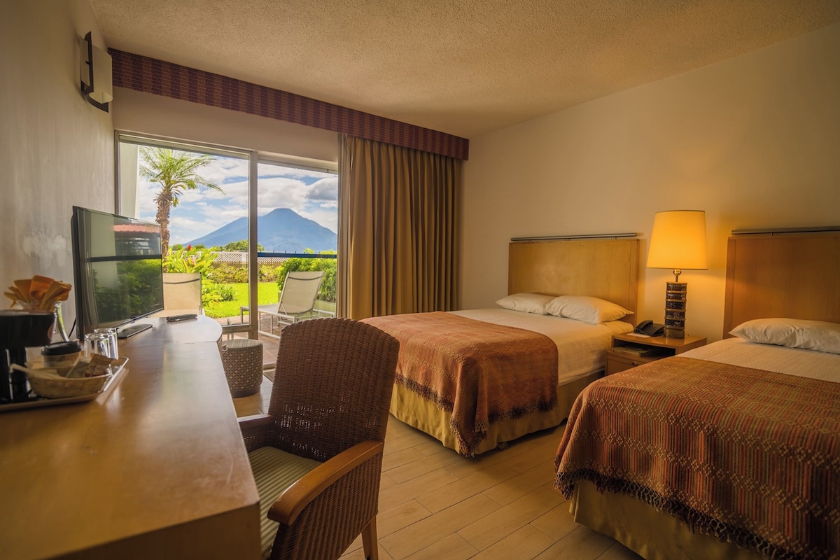 A hotel room with two beds, a desk with a television, and a view of a mountain.