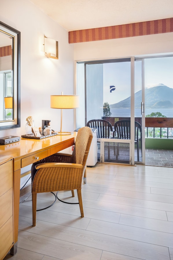 A hotel room with a wooden desk, a wicker chair, a lamp, and a view of mountains through the sliding glass door. - Porta Hotel del Lago (Panajachel)