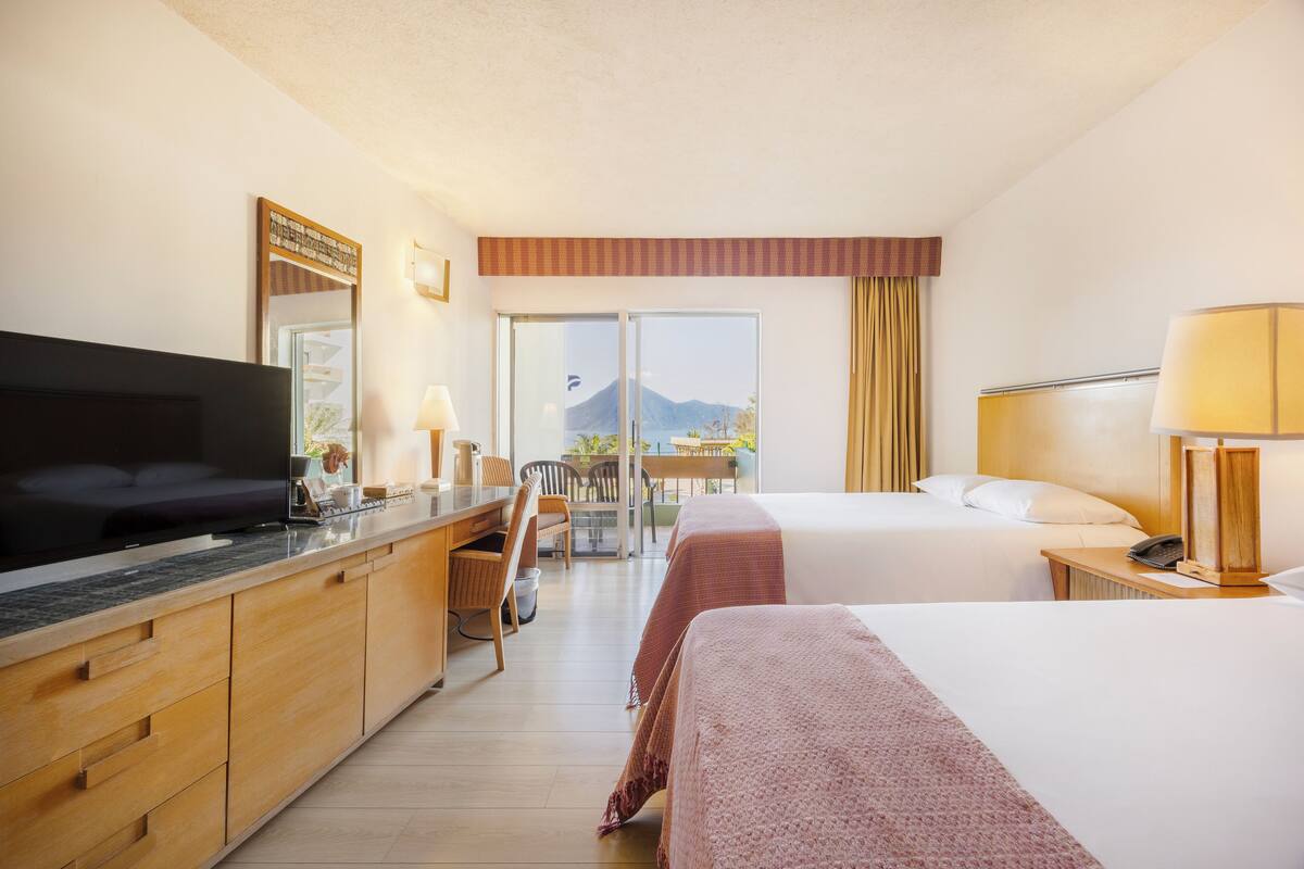 A hotel room with a TV, desk, two beds, a balcony with a mountain view, and a wooden cabinet.