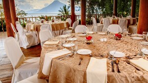 A dining area with tables set for a meal, overlooking a scenic view of a volcano and the sea. - Porta Hotel del Lago (Panajachel)