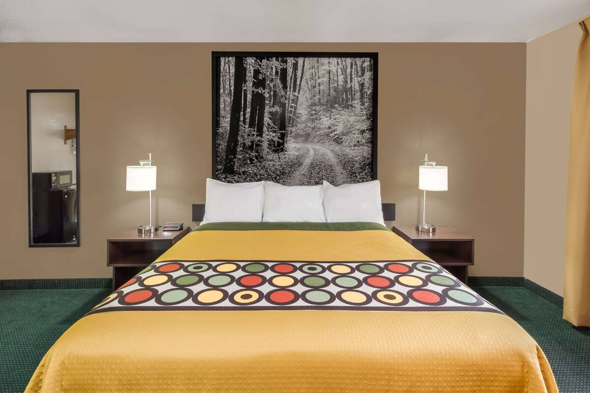 A hotel room with a large bed, two bedside tables, a nightstand, a mirror, and a wall-mounted picture of a forest path.