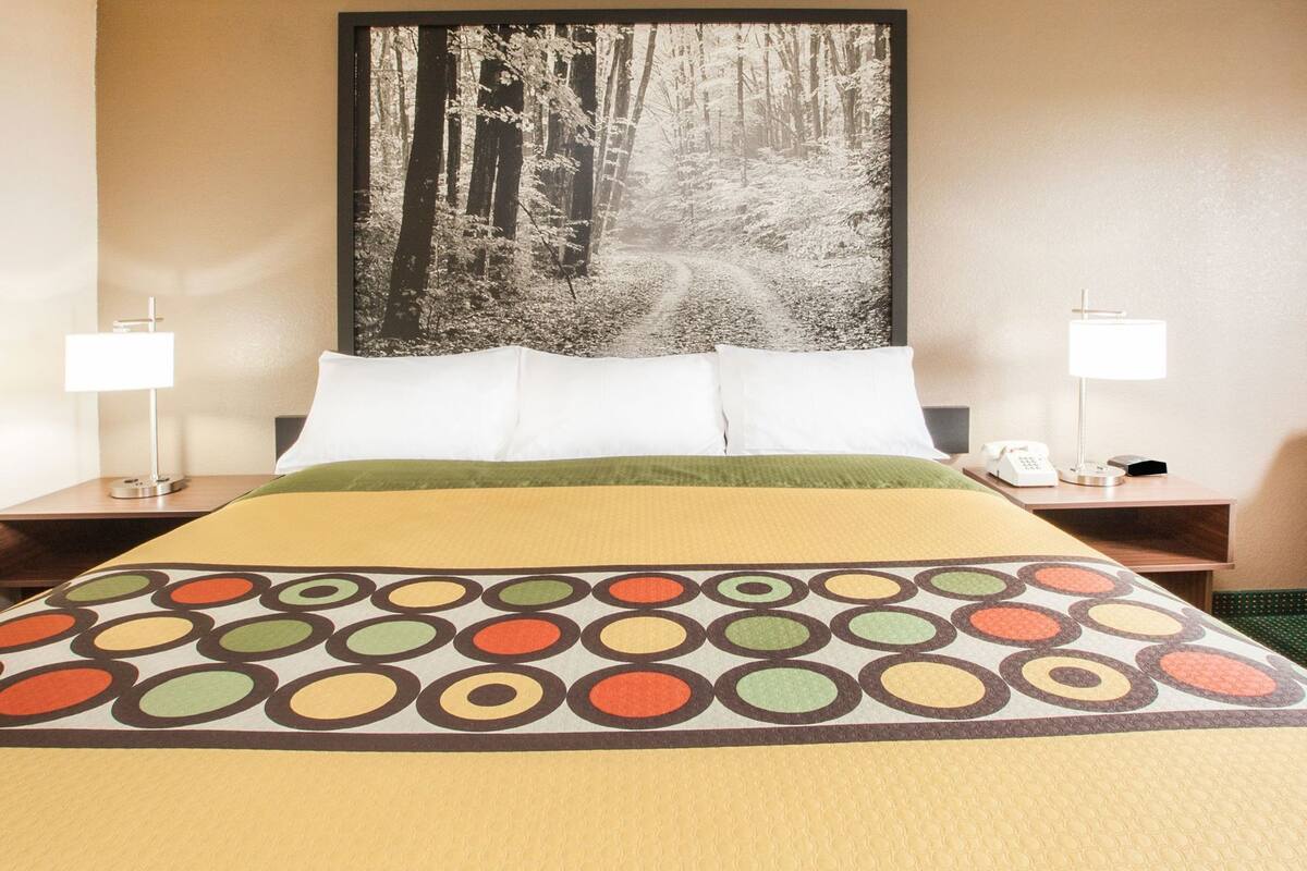 A hotel room with a bed, two bedside tables with lamps, a large forest-themed painting on the wall, and a colorful bedspread.