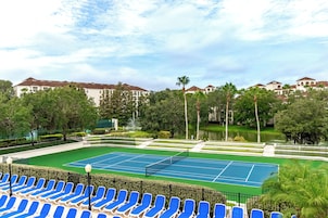 En tennisbana med blå stolar, omgiven av grönska och en vattenyta, med byggnader i bakgrunden.