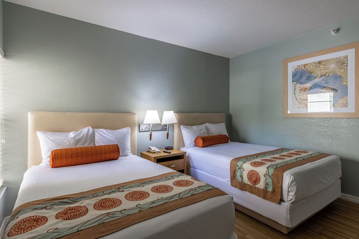 Habitación de hotel con dos camas, una mesita de noche con lámparas, un cuadro en la pared y una ventana con persianas.