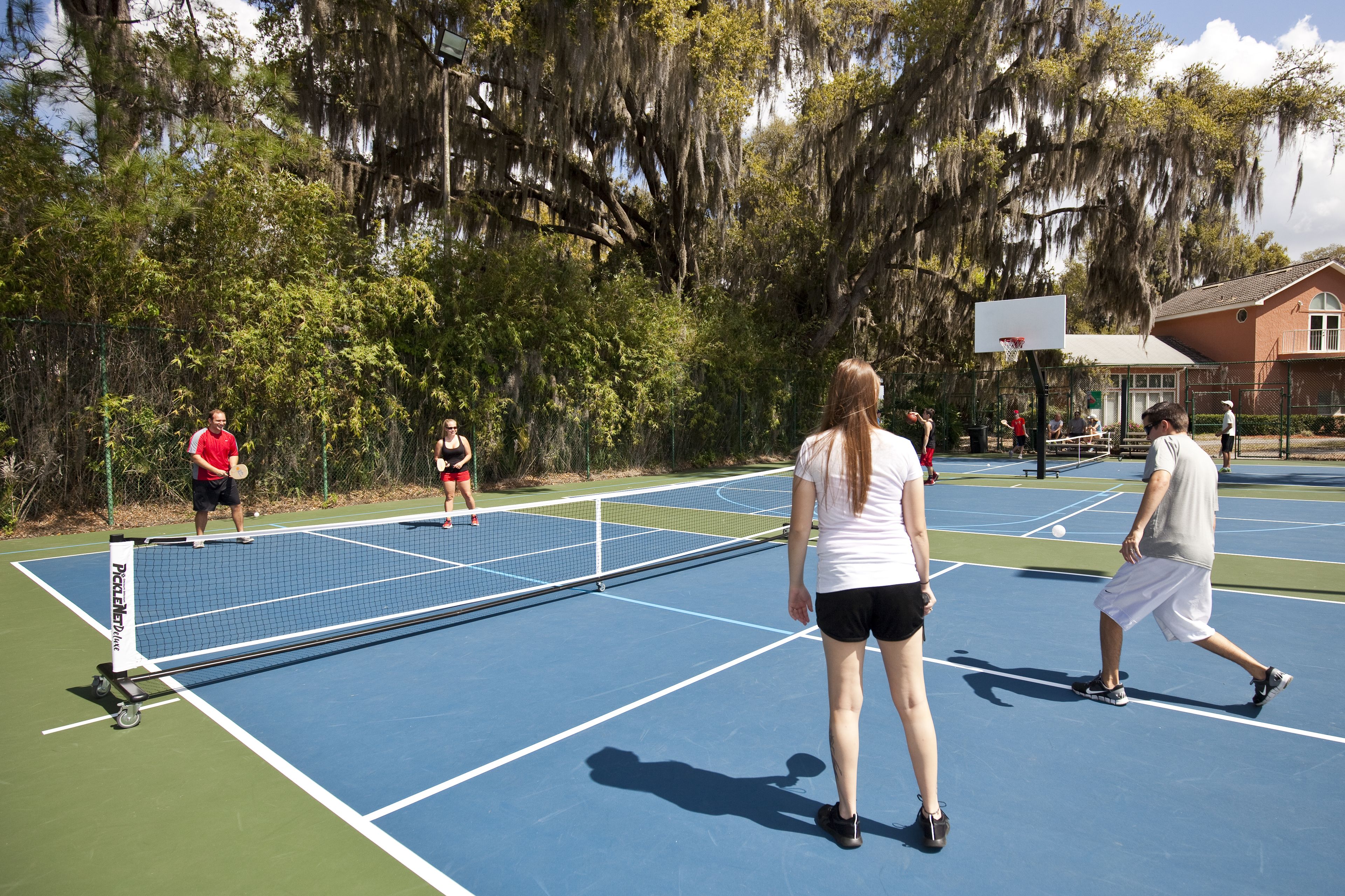 Una cancha de tenis al aire libre con red, rodeada de árboles y un edificio al fondo.
