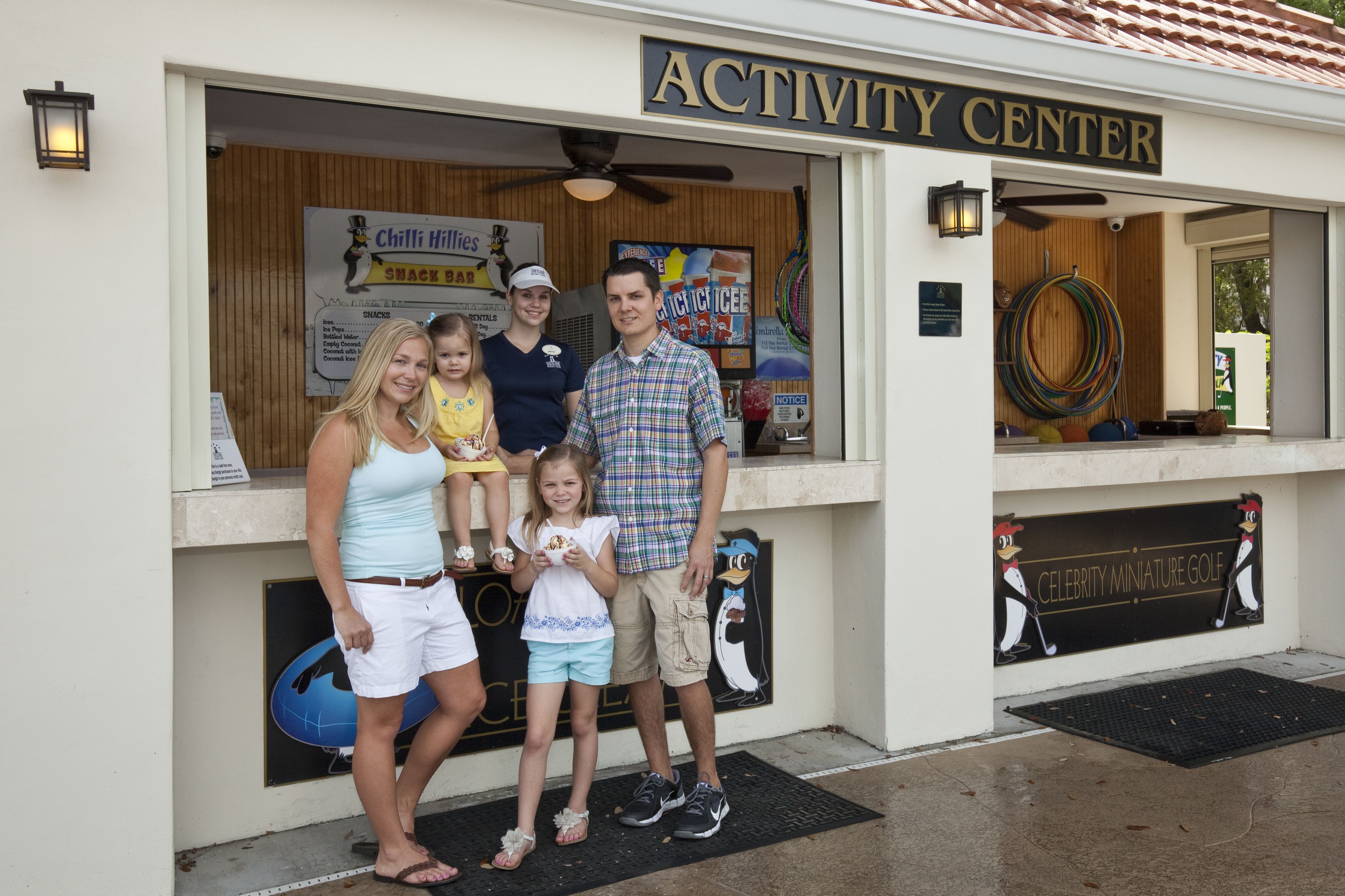 Eine Familie steht vor einem Aktivzentrum, an dem Schilder mit den Aufschriften 'ACTIVITY CENTER' und 'CELEBRITY MINIATURE GOLF' befestigt sind.