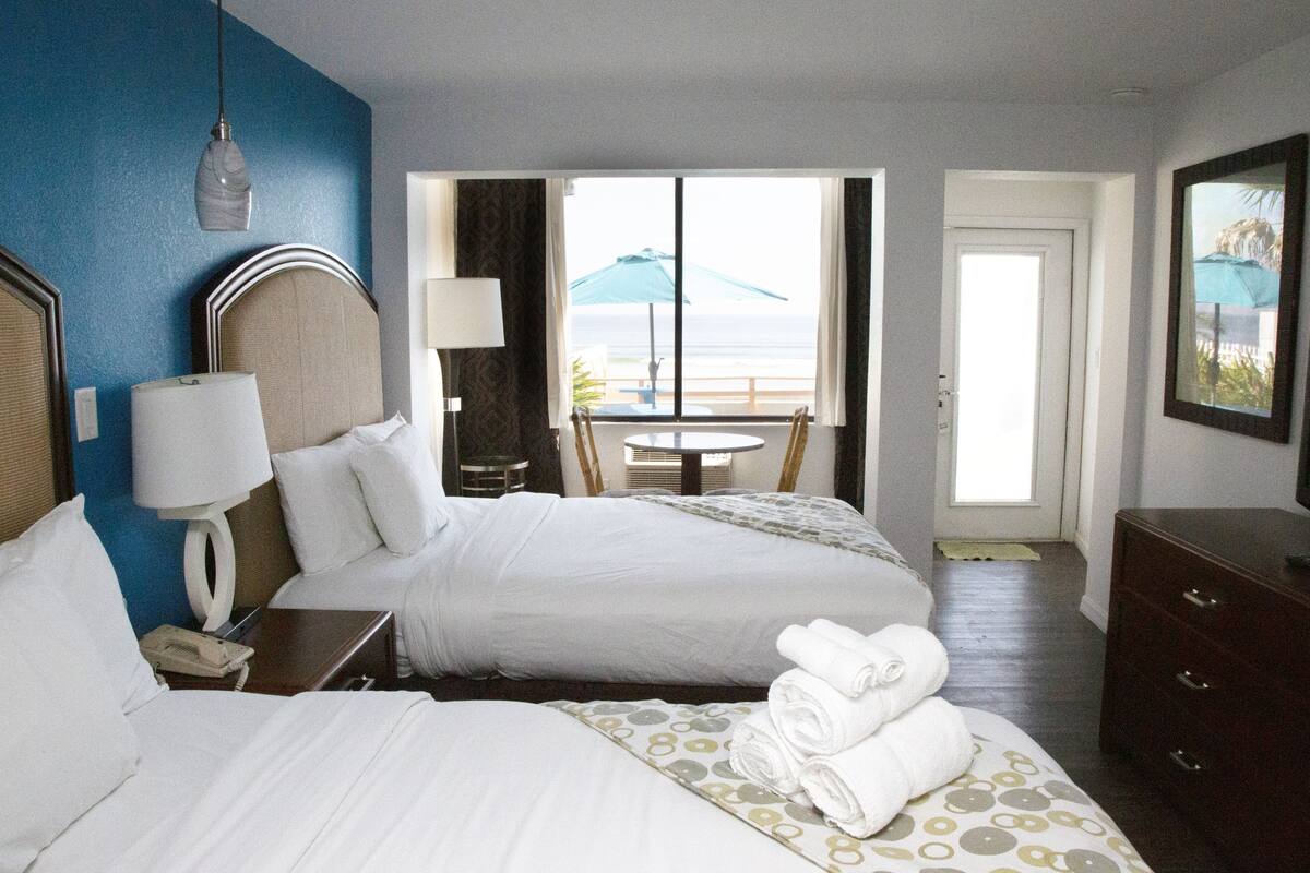 Quarto de hotel com duas camas, uma escrivaninha com abajur, cômoda e vista para o oceano através de uma porta.