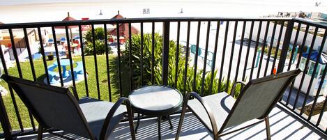 A balcony with two chairs and a small table, overlooking a beach with a pool and umbrellas.