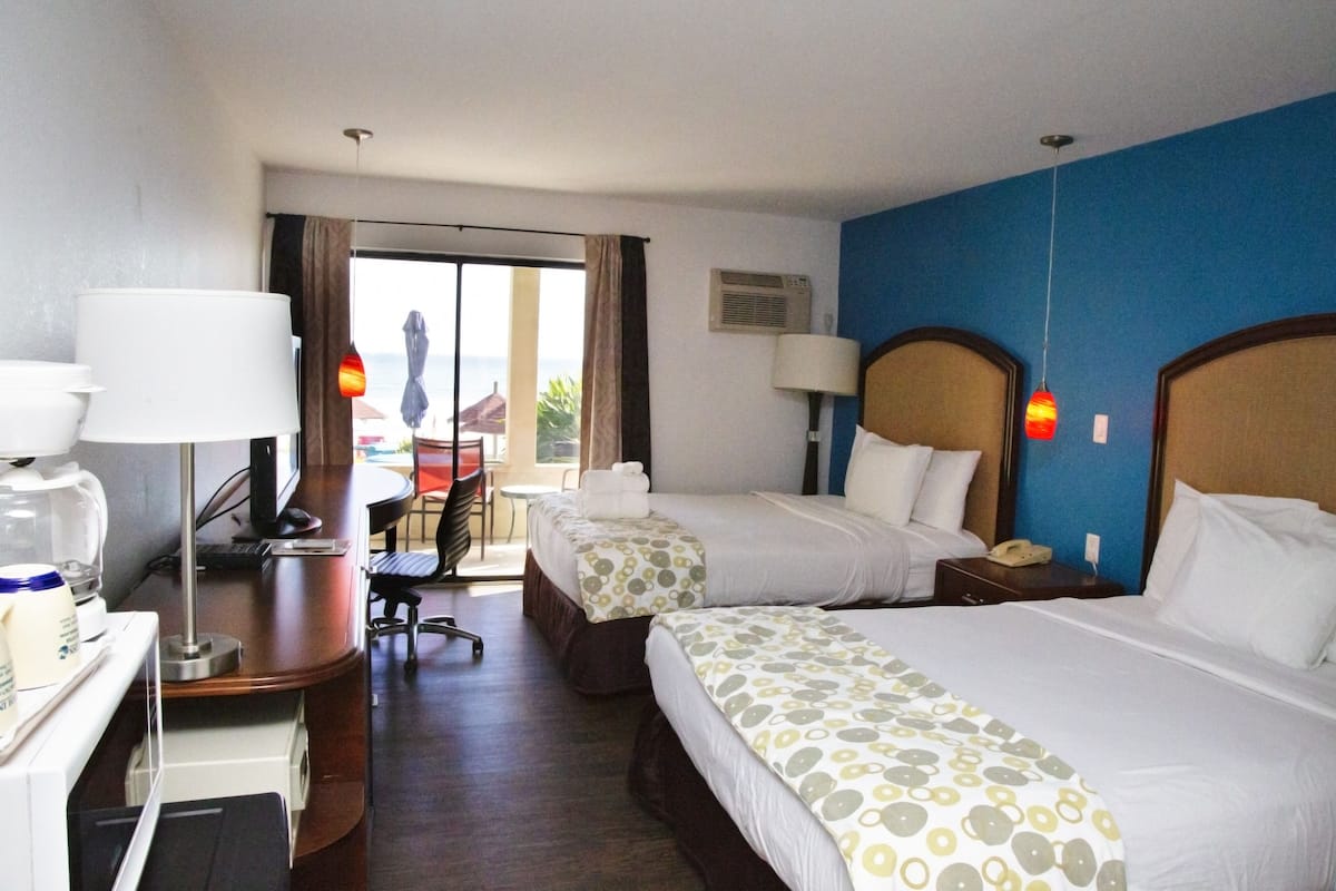 Quarto de hotel com duas camas, uma escrivaninha com computador, uma cadeira e vista para o exterior.