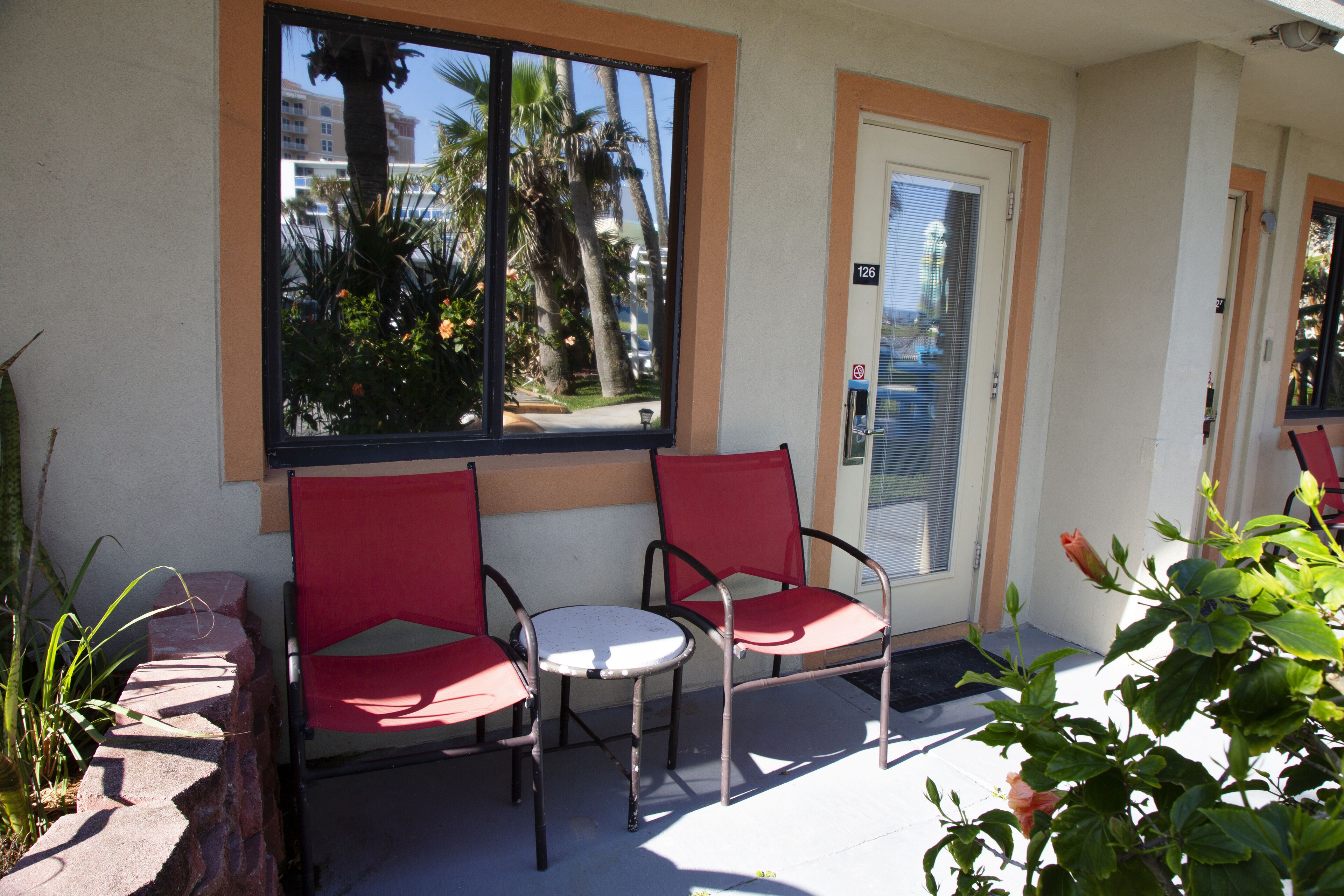 A patio area with two red chairs and a small table, a door with a number, and a view of palm trees and buildings.