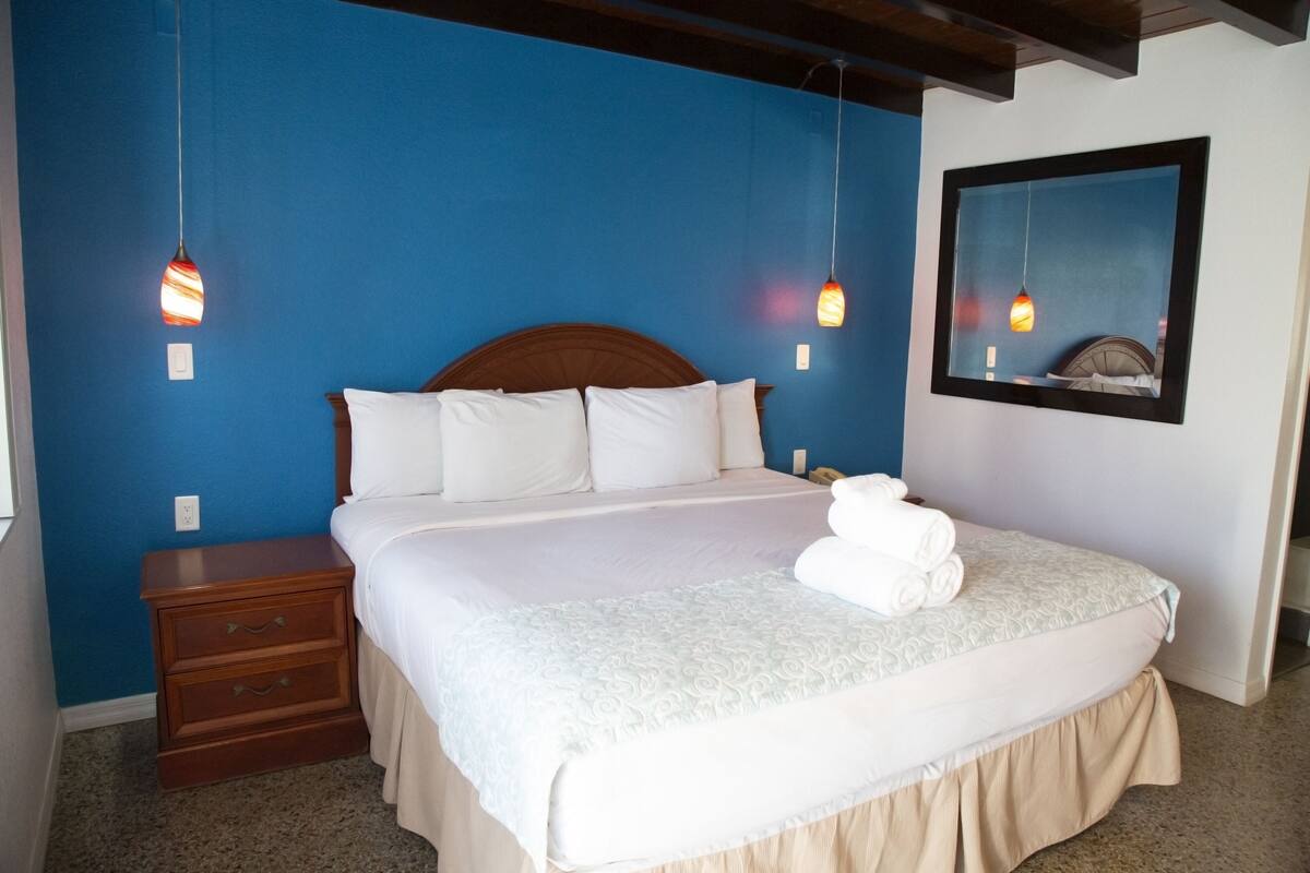 Uma cama bem arrumada com lençóis brancos e saia bege, uma mesa de cabeceira de madeira com gavetas e uma luminária pendente.