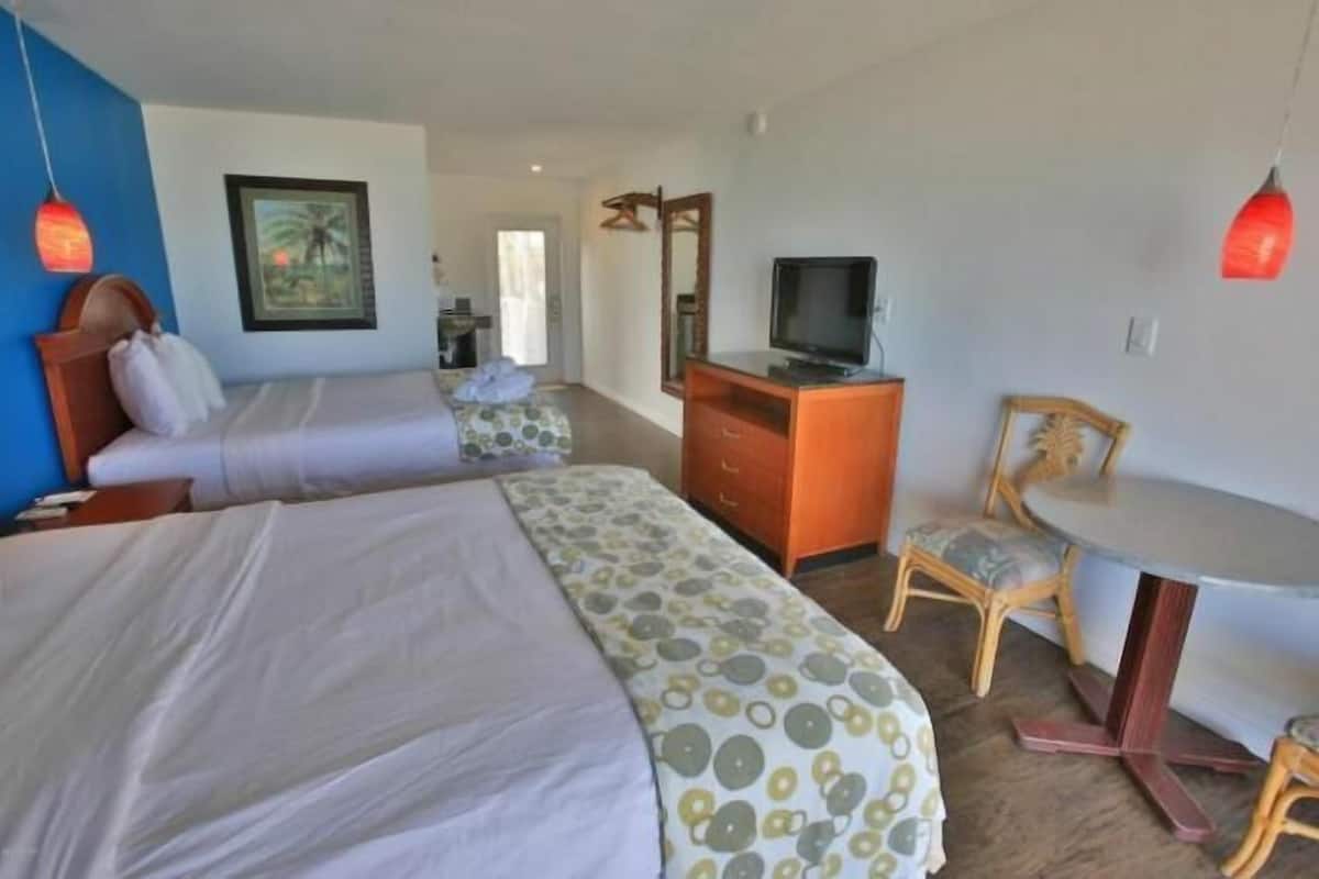 Quarto de hotel com duas camas, uma escrivaninha com televisão, uma cadeira e uma mesa de jantar.