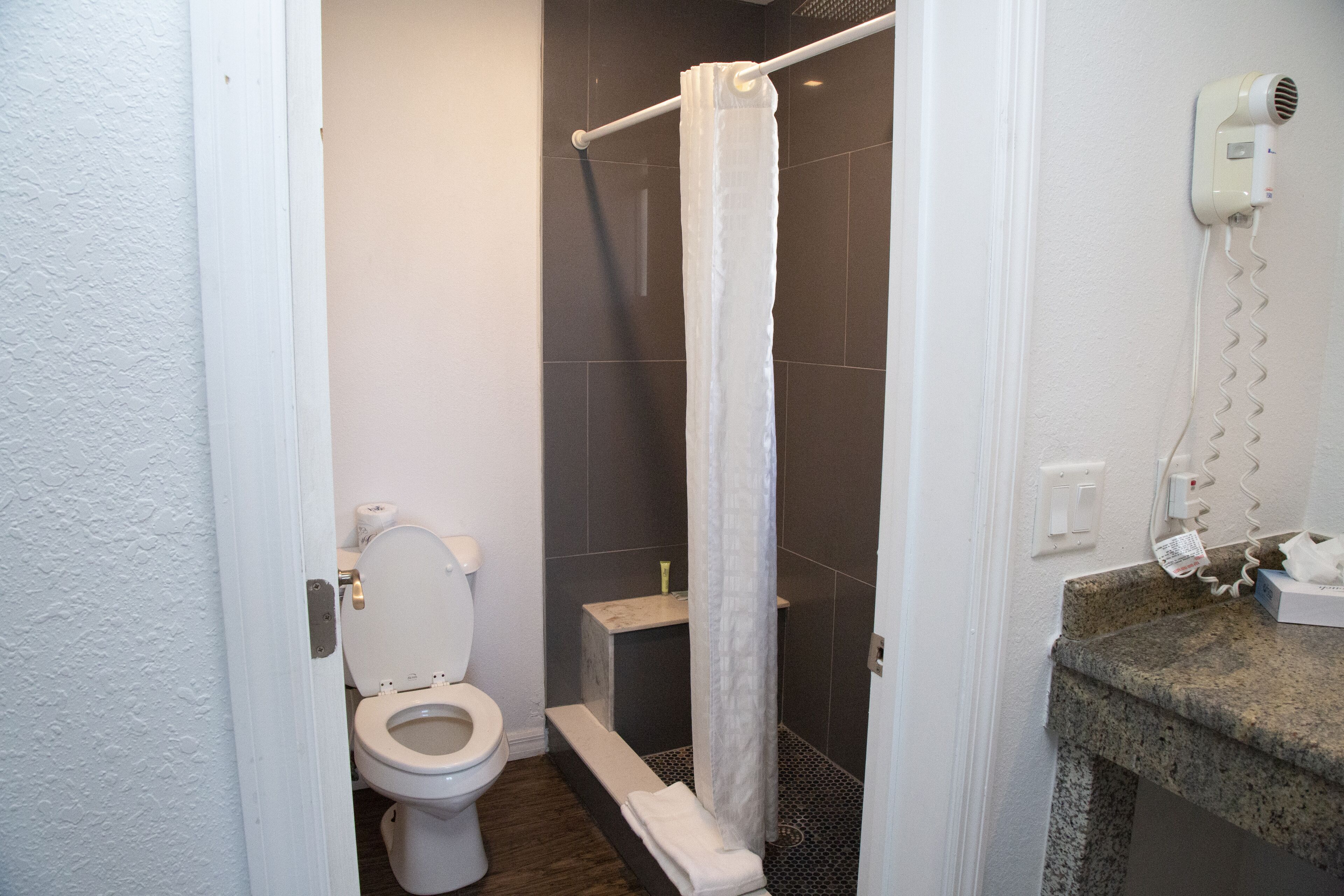A bathroom with a toilet, a shower with a white curtain, a hairdryer on the wall, and a granite countertop.