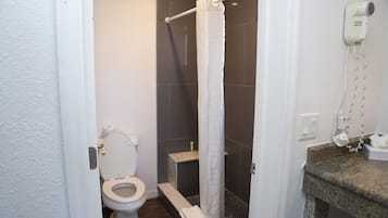 A bathroom with a toilet, a shower with a white curtain, a hairdryer on the wall, and a granite countertop.
