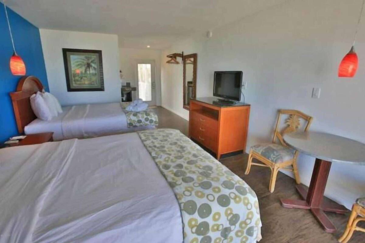 Quarto de hotel com duas camas, uma escrivaninha com televisão, uma cadeira e um quadro na parede.