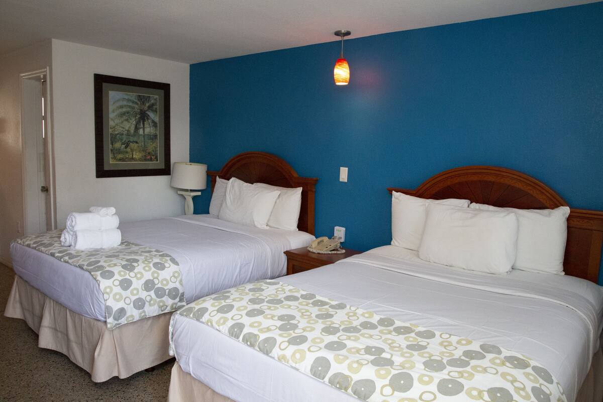 Quarto de hotel com duas camas, cada uma com cabeceira de madeira, roupa de cama branca e manta estampada. Há uma mesa de cabeceira com luminária e telefone em um dos lados.