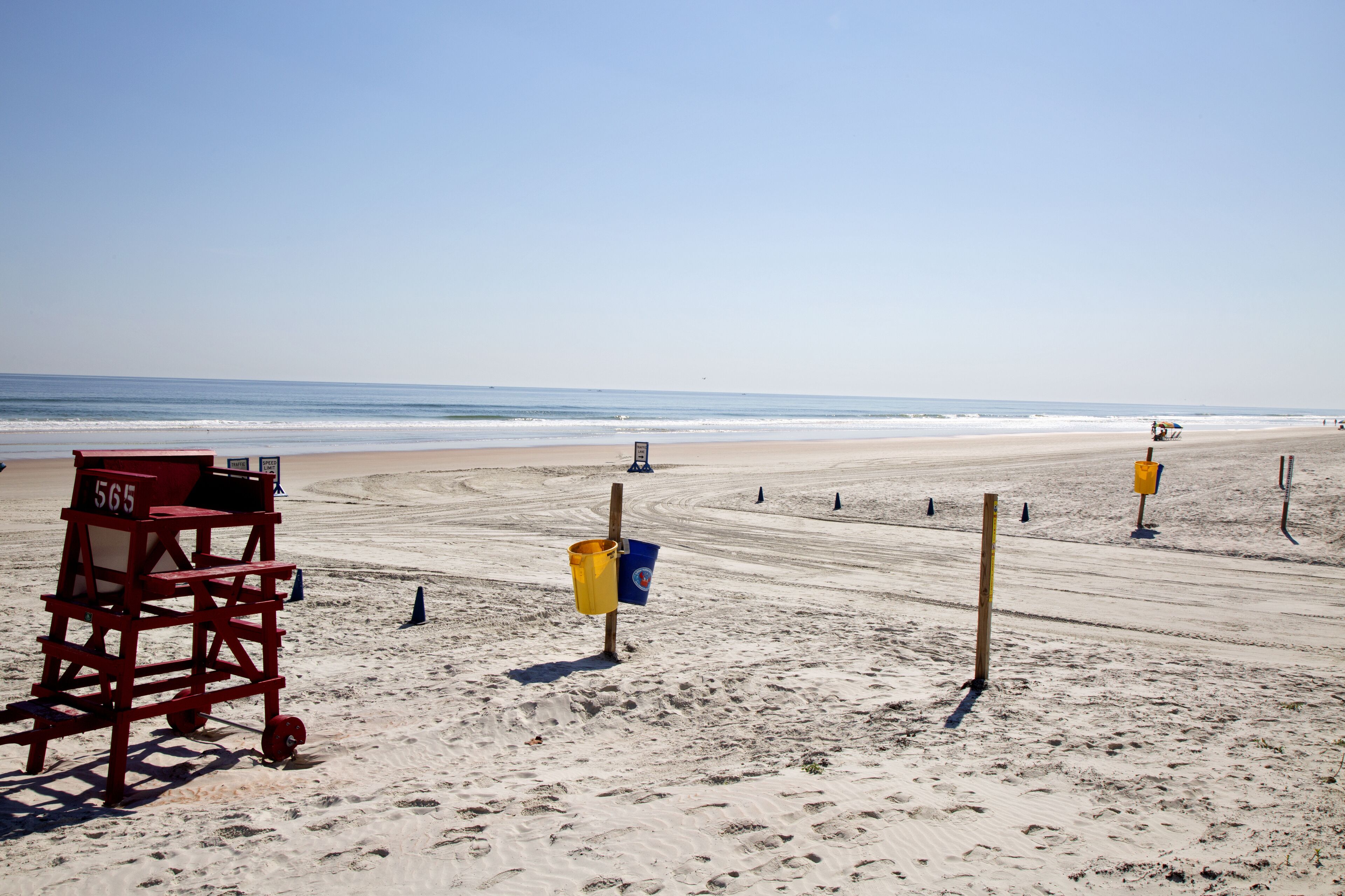 Una silla de socorrista con la etiqueta 565, un cubo amarillo y varios postes sobre una playa arenosa, con el océano al fondo.