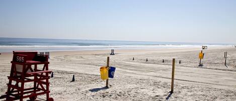 Una silla de socorrista con la etiqueta 565, un cubo amarillo y varios postes sobre una playa arenosa, con el océano al fondo.