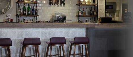 Zona de bar con taburetes, estanterías llenas de botellas, una máquina de café y un reloj en la pared.