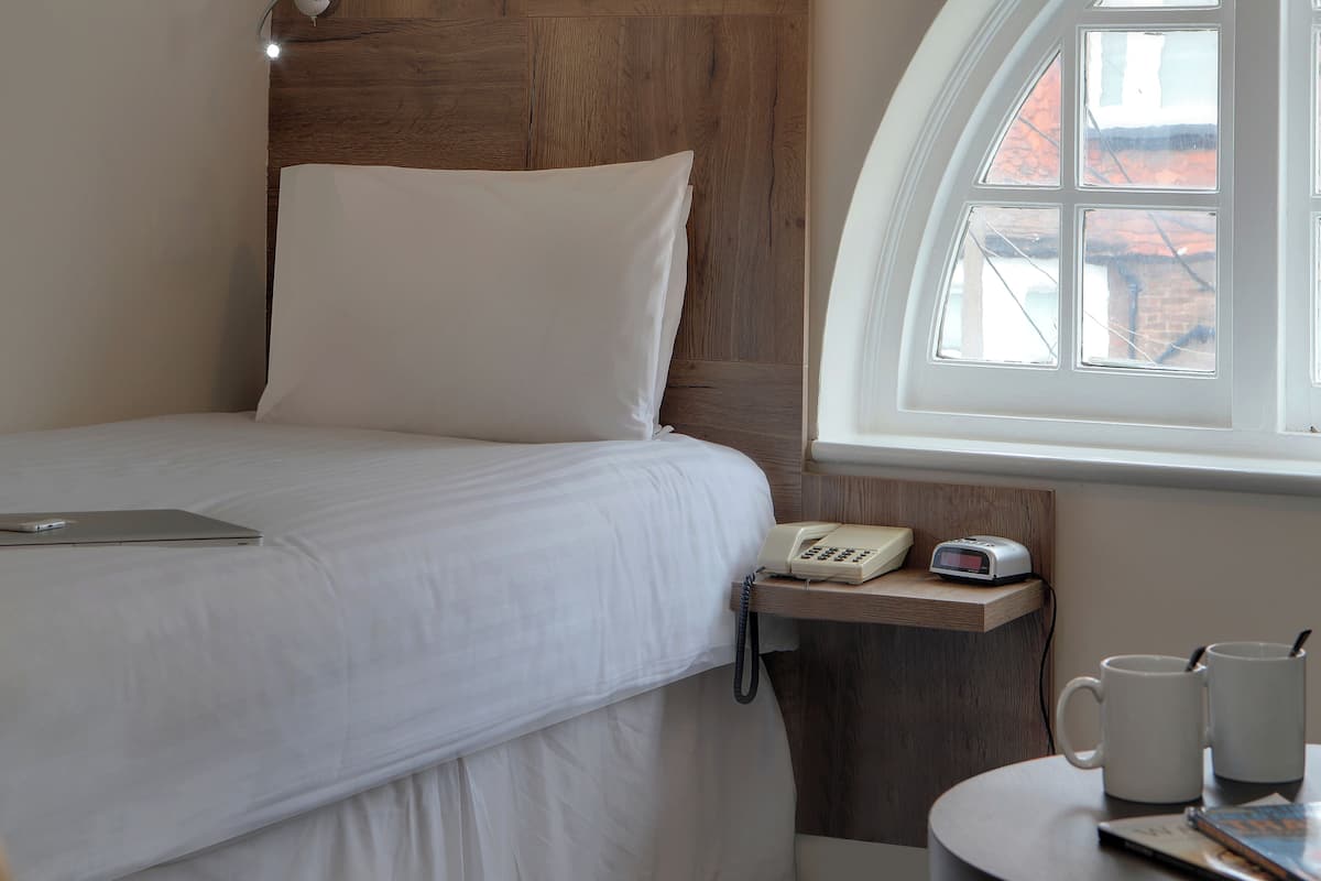 Een keurig opgemaakt bed met een laptop, een telefoon op het nachtkastje en een klok op een houten plank.