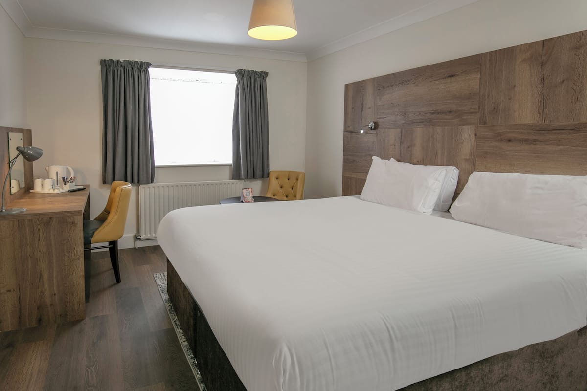 Een hotelkamer met een groot bed, een houten hoofdbord, een bureau met lamp, een stoel en een raam met gordijnen.