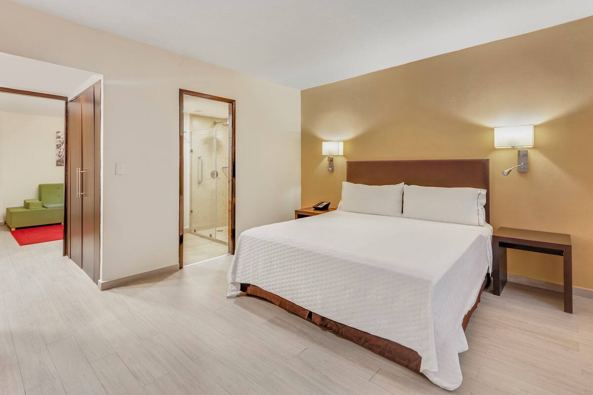 A hotel room with a large bed, bedside tables, a nightstand, a shower area, and a green chair.