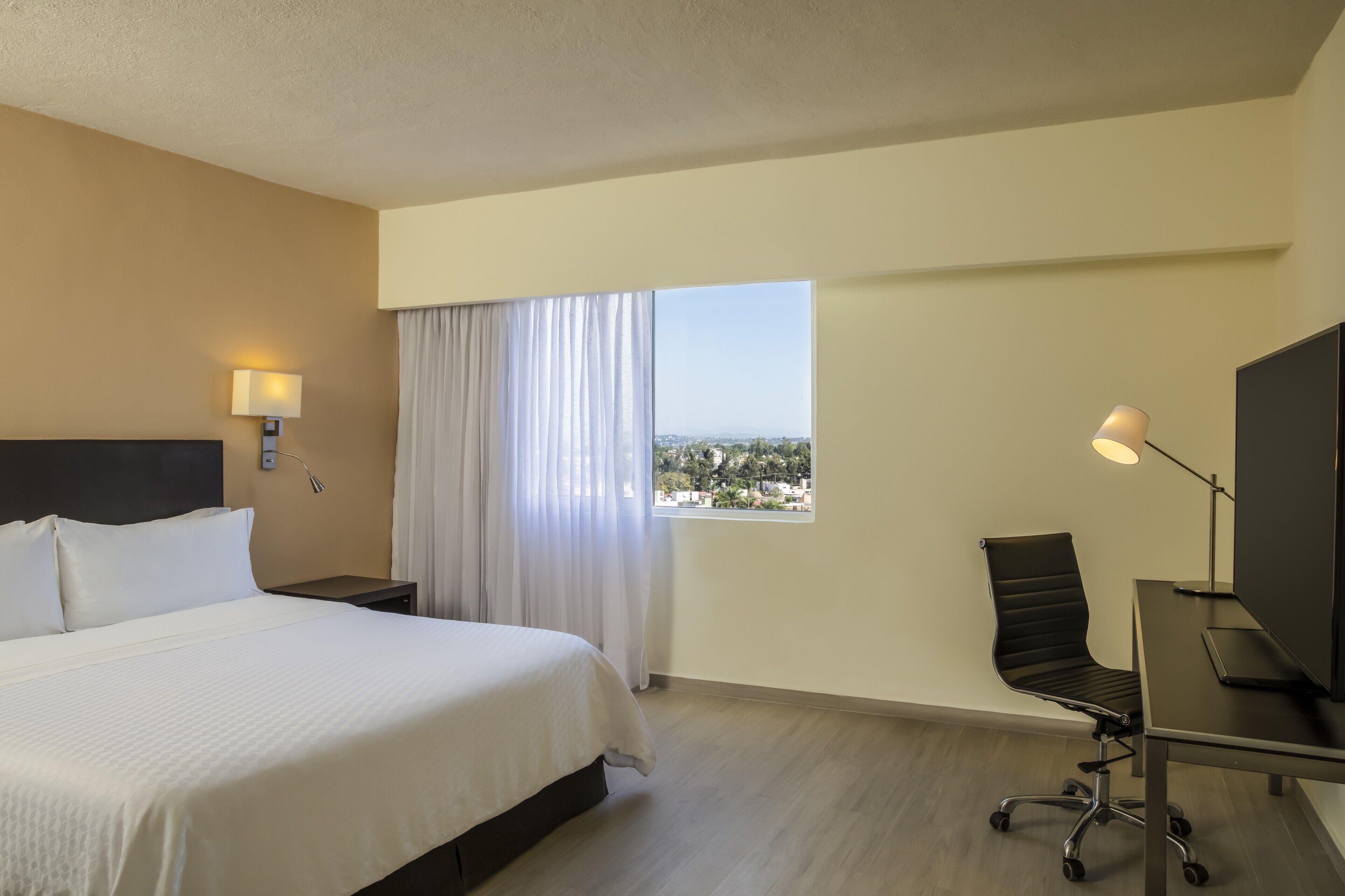 A hotel room with a bed, a desk with a computer, a chair, a TV, and a window with curtains.
