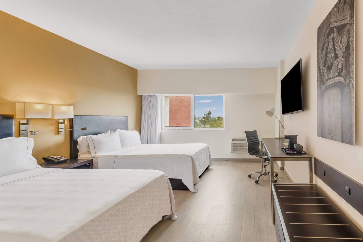 A hotel room with two beds, a desk, a chair, a TV, and a window with a view of the sky and trees.
