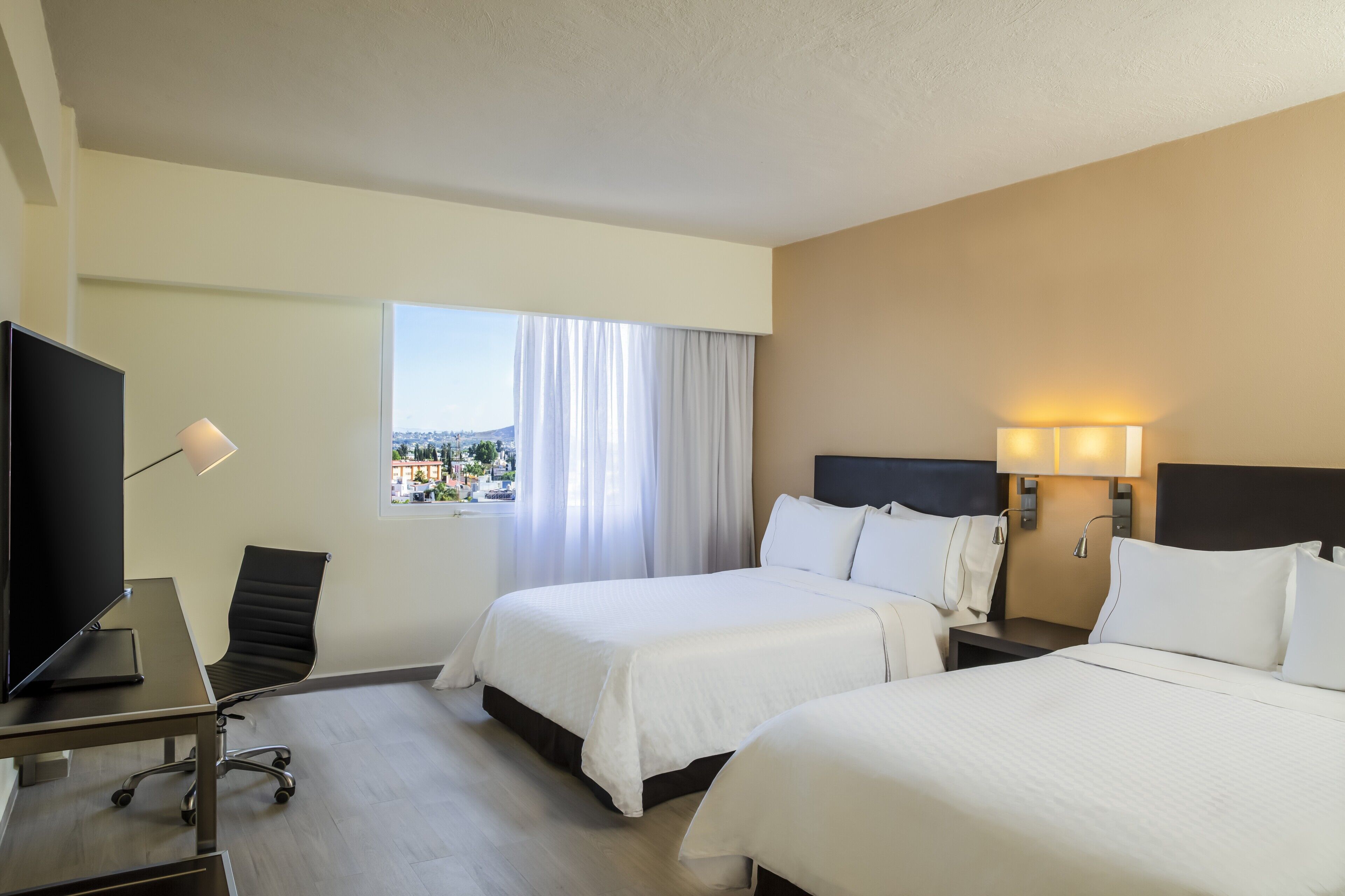 A hotel room with two beds, a desk with a computer, a chair, and a window with curtains.