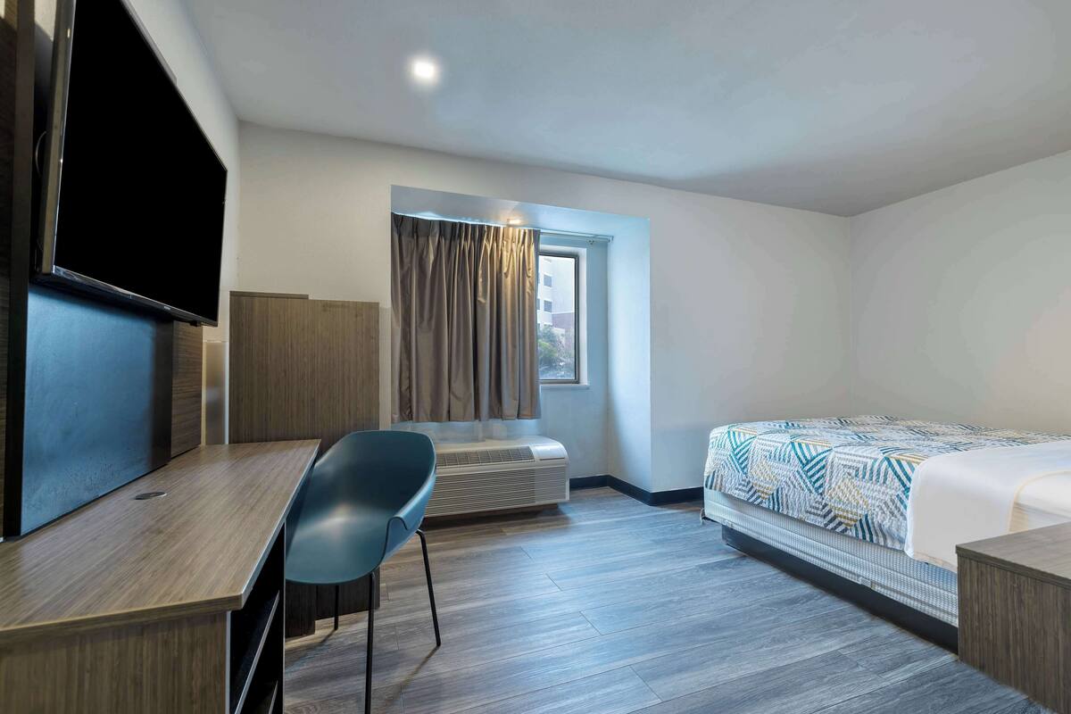 A hotel room with a bed, a desk, a chair, a TV, and a window with curtains.