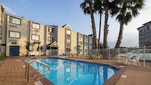 A swimming pool with a tiled deck, palm trees, and a building with multiple windows and balconies.