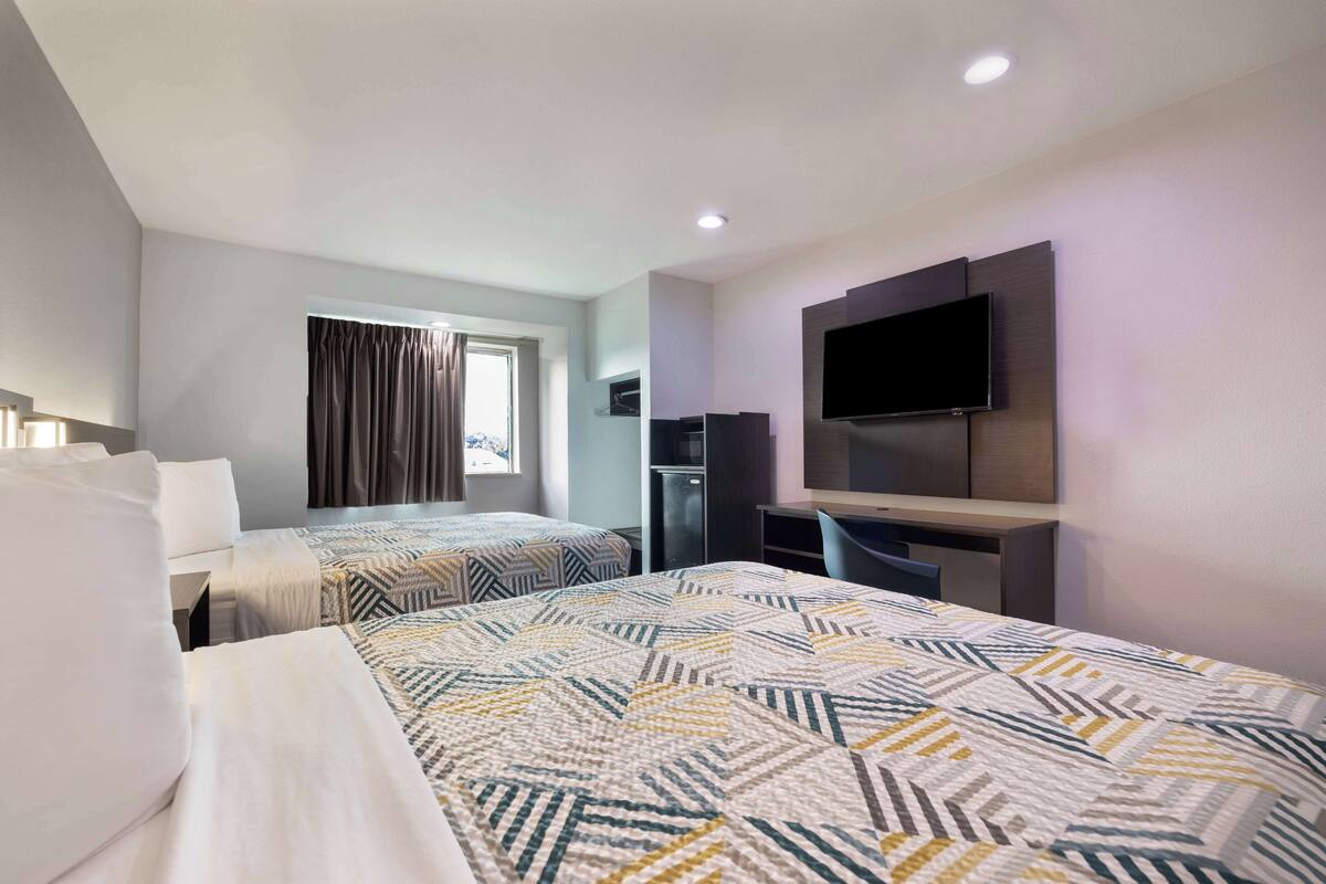A hotel room with two beds, a flat-screen TV, a desk, and a window with curtains.