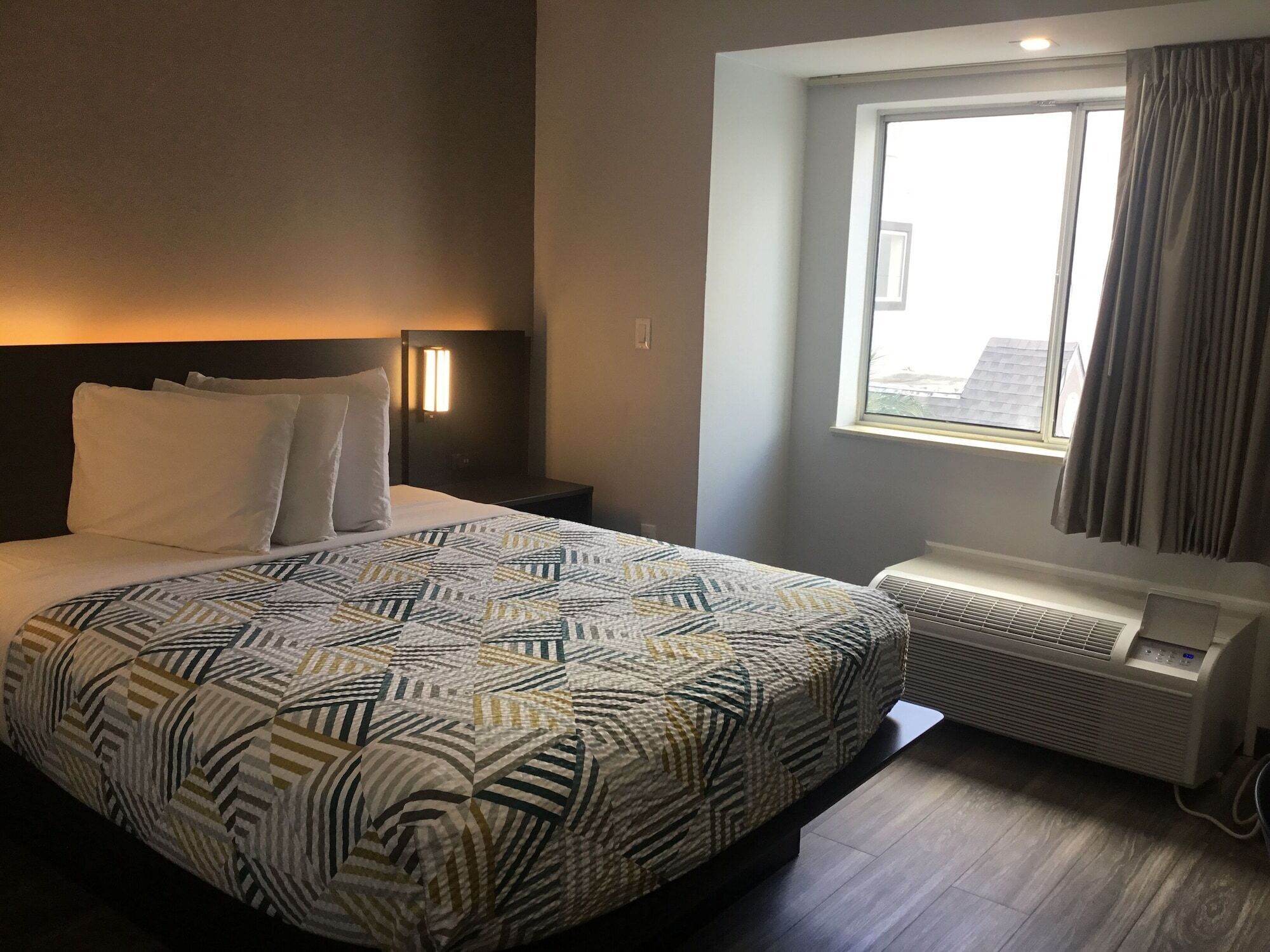 Ett hotellrum med en stor säng, ett nattduksbord, ett fönster med gardiner och en luftkonditioneringsanläggning.
