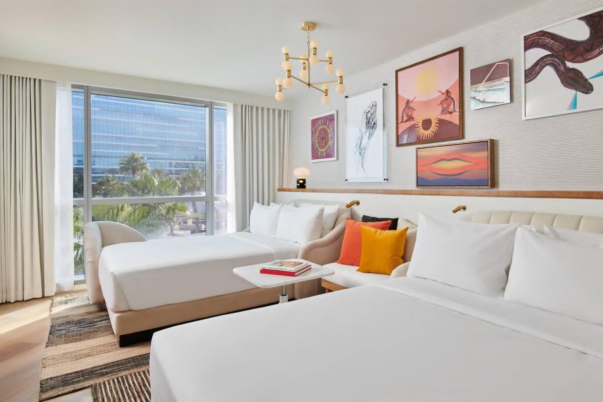 A hotel room with two beds, a large window with a view of palm trees, and artwork on the walls.
