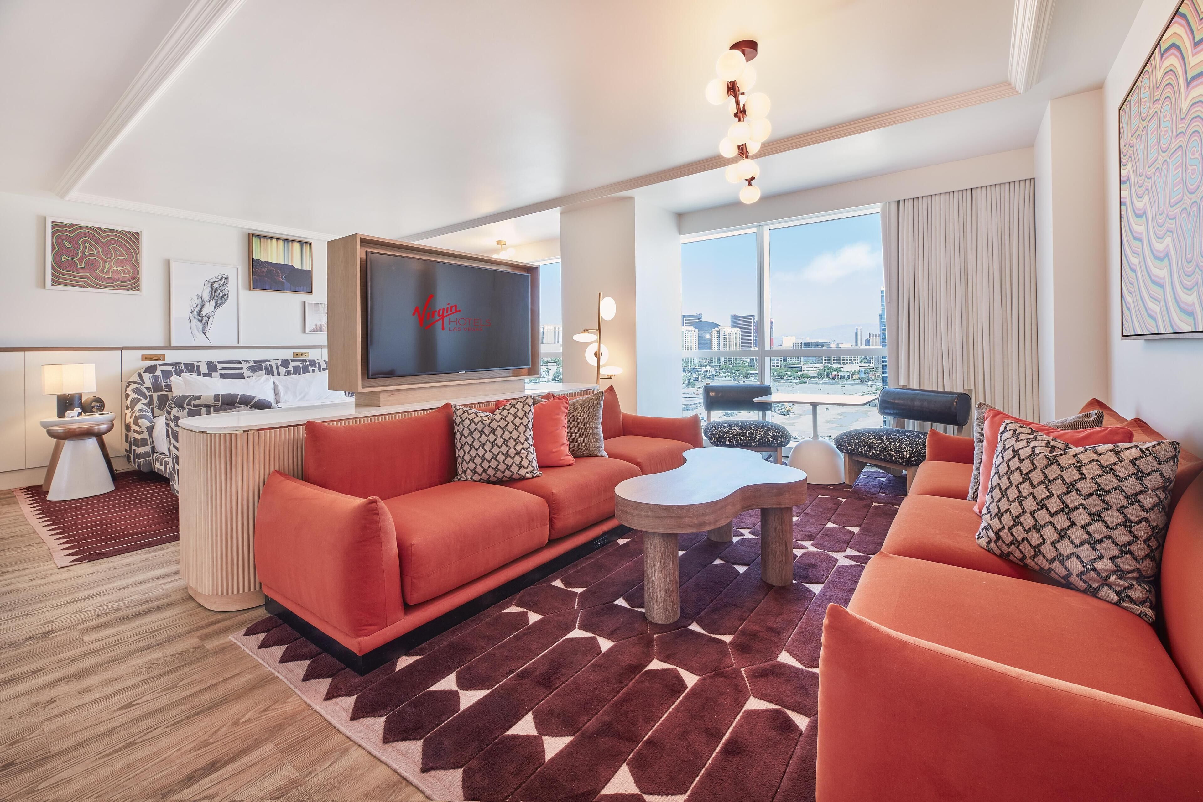 A modern hotel room with a red sofa set, a flat-screen TV, a dining area, and a city view.