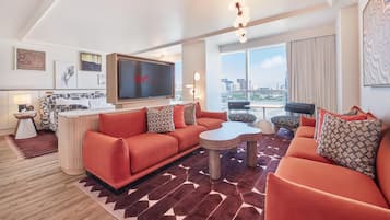 A modern hotel room with a red sofa set, a flat-screen TV, a dining area, and a city view.