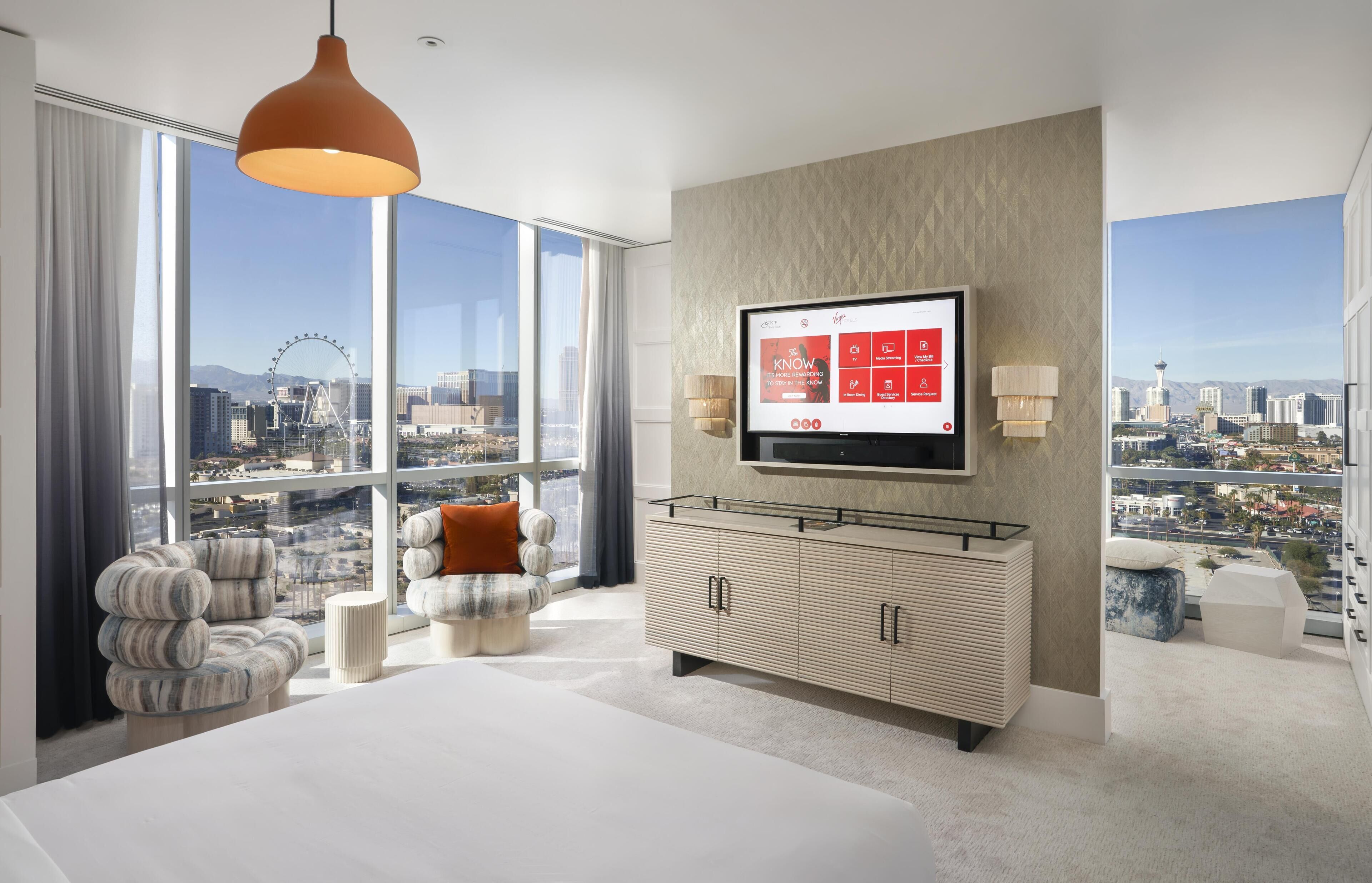 Una habitación de hotel moderna con una cama grande, un televisor montado en la pared, vistas a la ciudad y un ventanal amplio.