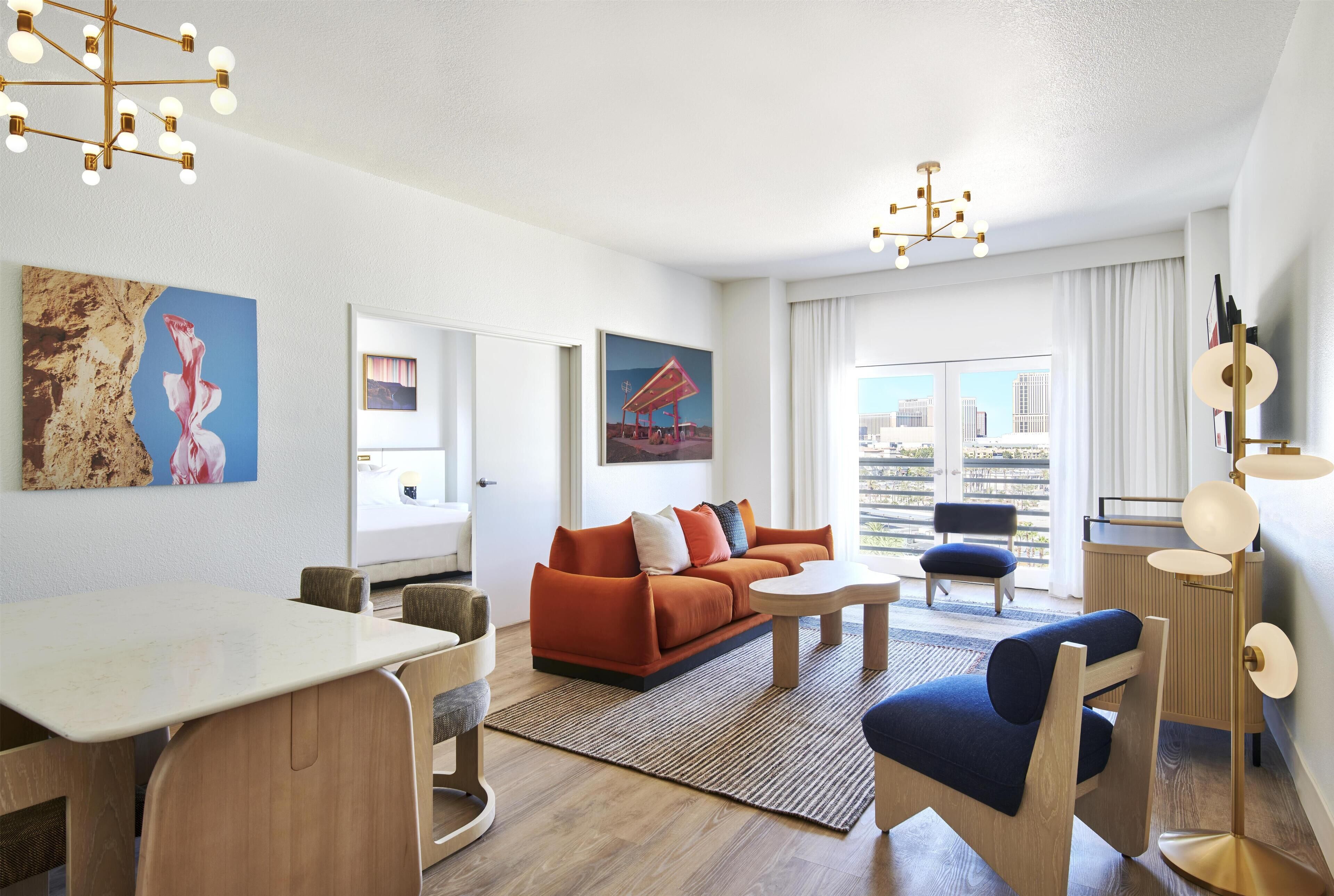 Una sala de estar moderna con un sofá, una mesa de centro y un comedor. Hay cuadros en las paredes y vistas de la ciudad a través de amplias ventanas.
