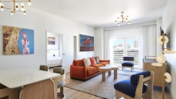 Una sala de estar moderna con un sofá, una mesa de centro y un comedor. Hay cuadros en las paredes y vistas de la ciudad a través de amplias ventanas.