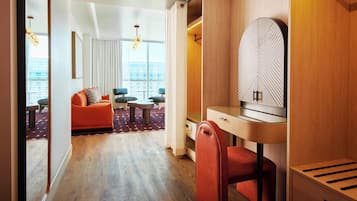 A modern hotel room with a wooden desk, a comfortable orange sofa, and a view of the city through large windows.