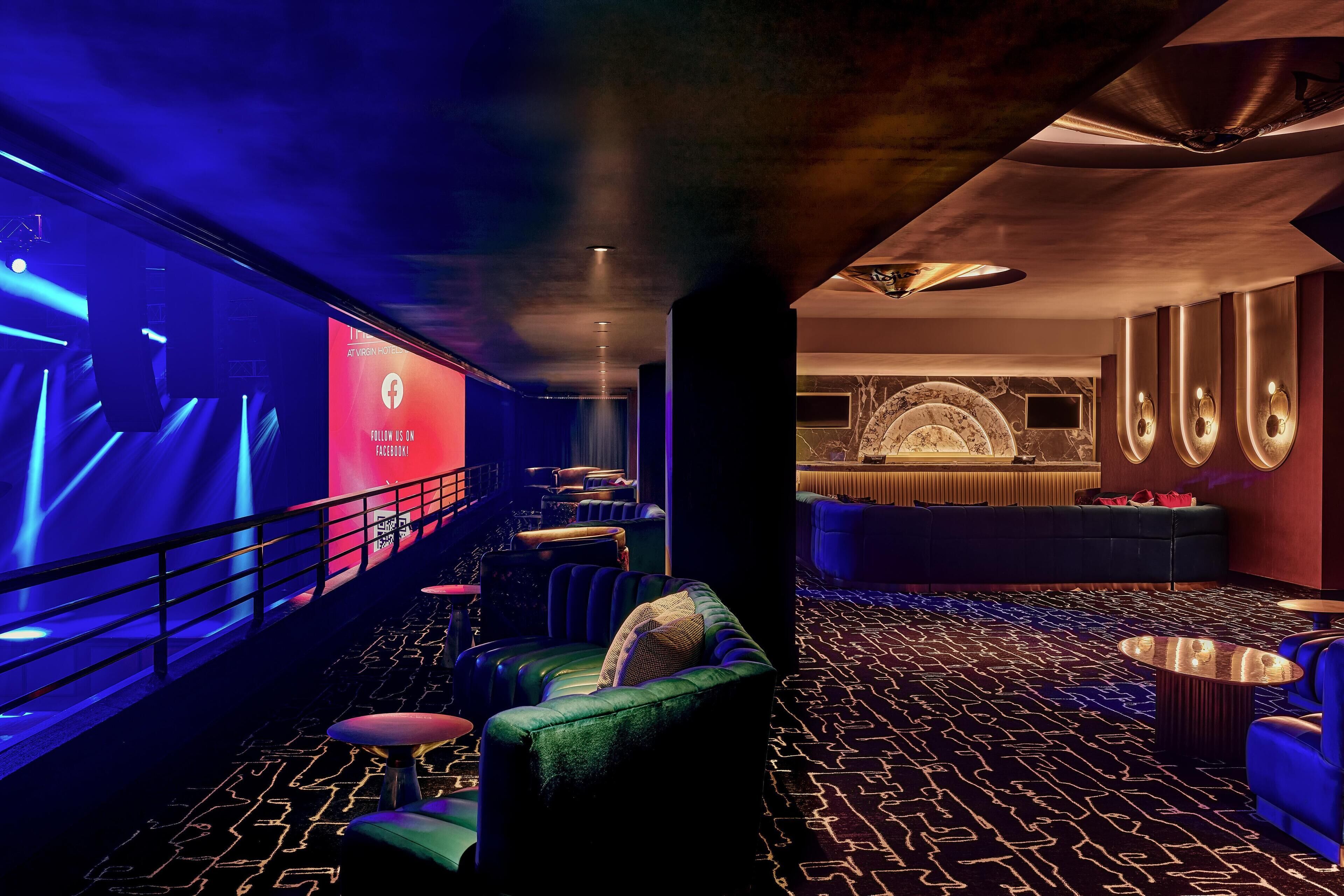 A modern lounge area with a plush green sofa, a bar area with a neon sign, and a patterned carpet.